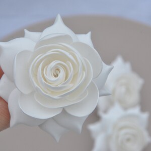 May include: Three white paper roses, one held in a hand, with the other two in the background.