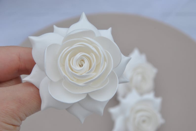 May include: Three white paper roses, one held in a hand, with the other two in the background.