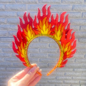 May include: A red and yellow felt headband with a flame design. The headband is made of a flexible material and is decorated with glitter.