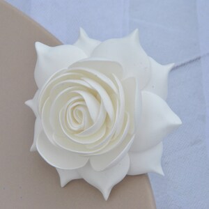 May include: A single white artificial rose with a layered, curled center. The rose is on a light brown surface.