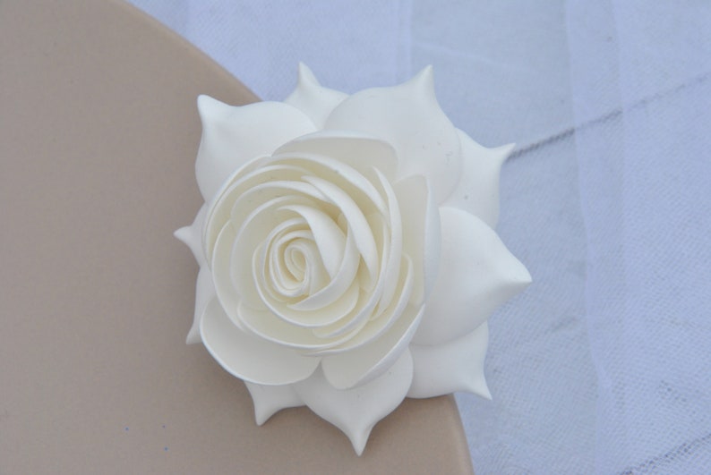 May include: A single white artificial rose with a layered, curled center. The rose is on a light brown surface.