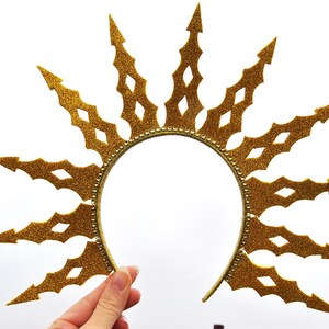 Puede incluir: Una diadema de purpurina dorada con un dise&ntilde;o de sol. La diadema est&aacute; hecha de un material flexible y tiene una forma puntiaguda en forma de estrella.