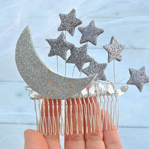 May include: Silver glitter crescent moon and star hair comb. The comb has a silver glitter finish and features a crescent moon with seven stars.