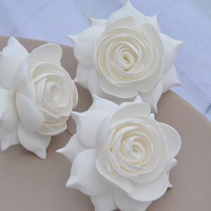 May include: Three white foam roses, each with a layered, curled center, are arranged on a light brown surface.