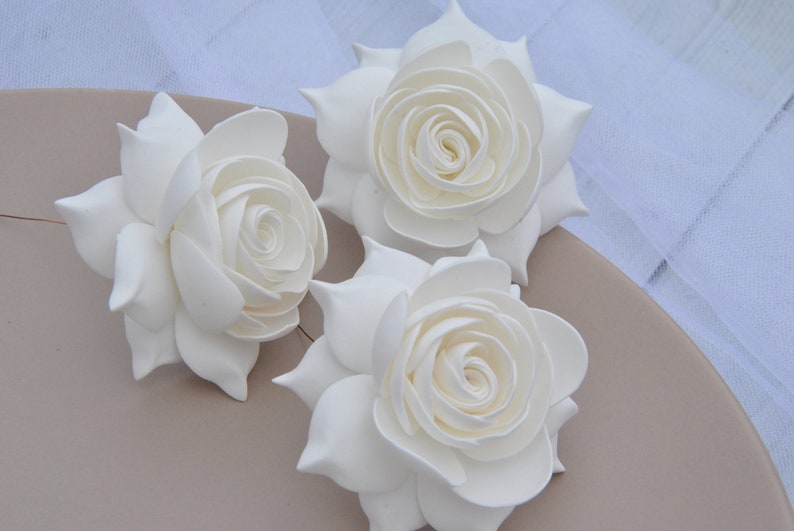 May include: Three white foam roses, each with a layered, curled center, are arranged on a light brown surface.