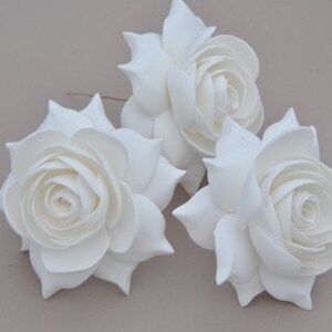 May include: Three white foam roses with a layered, ruffled design. The roses are arranged in a cluster on a light brown background.