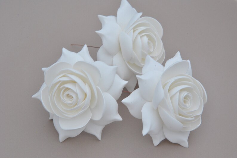 May include: Three white foam roses with a layered, ruffled design. The roses are arranged in a cluster on a light brown background.