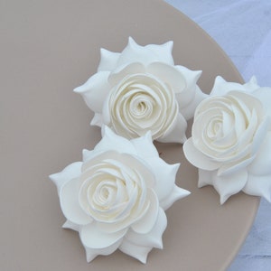 May include: Three white paper roses, each with a layered, curled center, are arranged on a light brown surface.