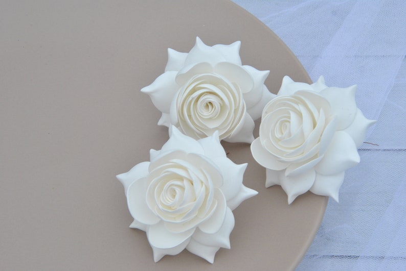 May include: Three white paper roses, each with a layered, curled center, are arranged on a light brown surface.