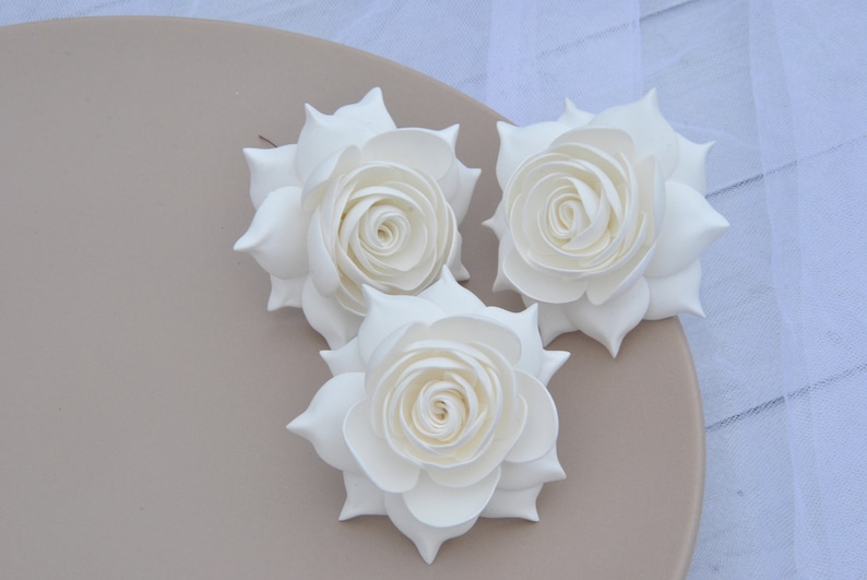 May include: Three white foam roses with a layered, ruffled design. The roses are arranged on a light brown plate.