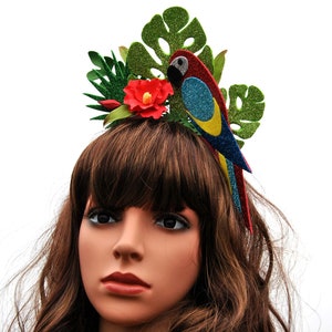 K&ouml;nnte beinhalten: Ein gr&uuml;nes, rotes, blaues und gelbes Glitzer-Papageien-Haarband mit einer roten Blume. Das Haarband wird von einer Puppe mit braunen Haaren getragen.