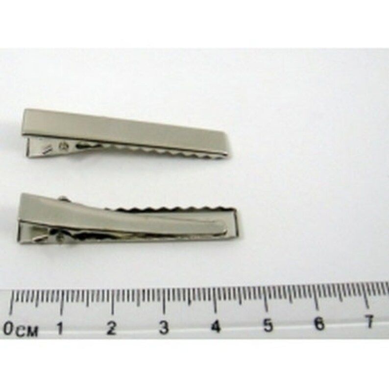 Puede incluir: Dos pinzas de cocodrilo plateadas, cada una mide aproximadamente 4 cm de largo, se muestran sobre un fondo blanco. Una regla es visible en la parte inferior de la imagen.