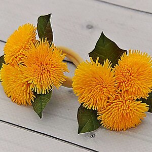 Puede incluir: Dos juegos de anillos de servilleta de flores de diente de le&oacute;n amarillas con hojas verdes. Los anillos est&aacute;n hechos de tela y est&aacute;n atados con una cinta marr&oacute;n.