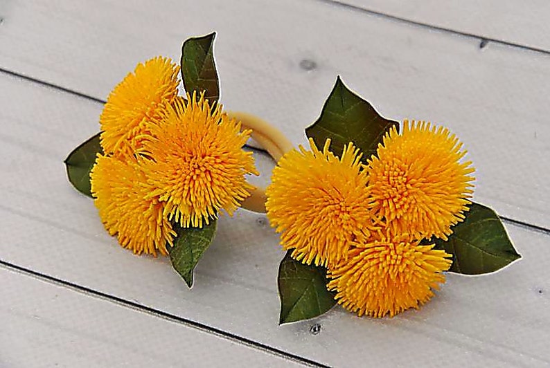 Puede incluir: Dos juegos de anillos de servilleta de flores de diente de le&oacute;n amarillas con hojas verdes. Los anillos est&aacute;n hechos de tela y est&aacute;n atados con una cinta marr&oacute;n.