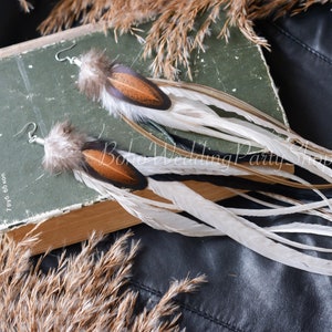 May include: A pair of long feather earrings with brown, white, and black feathers. The earrings are hanging from a book with the text "Boho Wedding Party Shop".