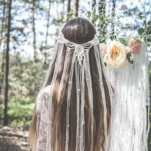 May include: A woman with long brown hair wearing a white crocheted headpiece with a floral design. The headpiece has long strands of white yarn hanging down her back. She is standing in a forest with a dreamcatcher behind her.