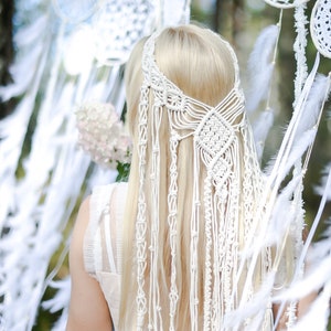 May include: A white macrame hairpiece with long strands hanging down the back. The hairpiece is worn by a person with long blonde hair. The hairpiece is in front of a background of white dreamcatchers with feathers.