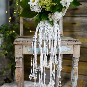 May include: A white macrame plant hanger with a white ceramic vase filled with white and green flowers. The vase is sitting on a rustic wooden table with a light blue painted surface.