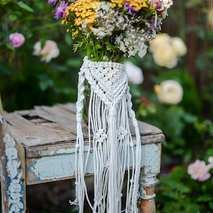 Op de afbeelding: Een boeket gele, witte en paarse wilde bloemen in een glazen pot met een witte macrame plantenhanger. De pot staat op een rustieke houten stoel met blauwe verf.