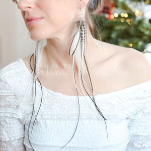 May include: Long feather earrings with white, black, and gray feathers. The earrings are hanging from a silver hook. The model is wearing a white off-the-shoulder top. The background is blurred.