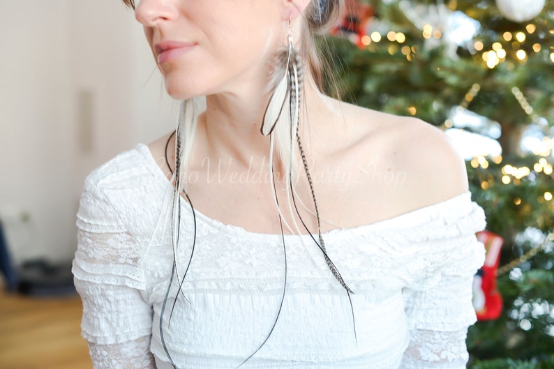 May include: Long feather earrings with white, black, and gray feathers. The earrings are hanging from a silver hook. The model is wearing a white off-the-shoulder top. The background is blurred.