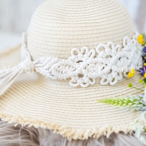 Puede incluir: Un sombrero de paja blanco con un borde y un borla de macramé blanco. El sombrero está sobre una superficie blanca peluda con un ramo de flores silvestres al lado.