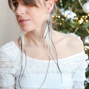 May include: Long, dangling earrings featuring white and black feathers. The earrings are suspended from silver-colored hooks. The model is wearing a white off-the-shoulder top. The background is a blurred Christmas tree.
