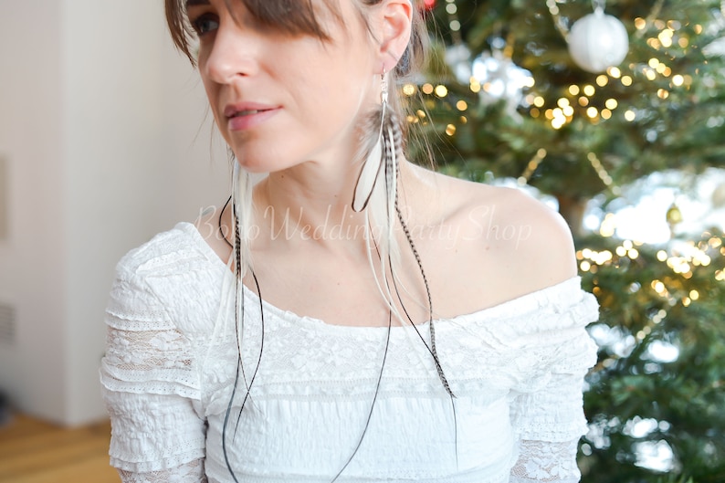 May include: Long, dangling earrings featuring white and black feathers. The earrings are suspended from silver-colored hooks. The model is wearing a white off-the-shoulder top. The background is a blurred Christmas tree.