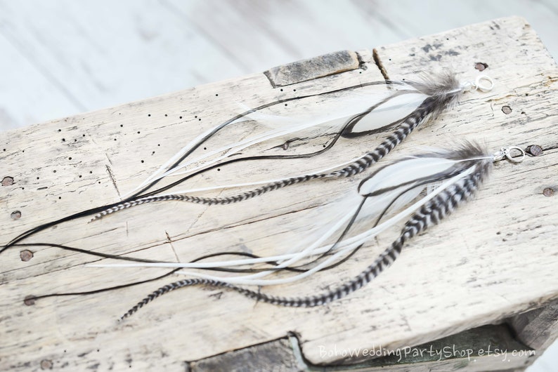 May include: Two feather earrings with white, black, and gray feathers. The earrings have a silver-colored clasp. The feathers are arranged on a weathered wooden surface, creating a bohemian aesthetic.