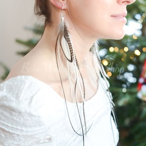 May include: Long feather earrings with a mix of white, brown, and black feathers. The earrings are suspended from silver hooks and dangle gracefully. The model is wearing a white off-the-shoulder top.