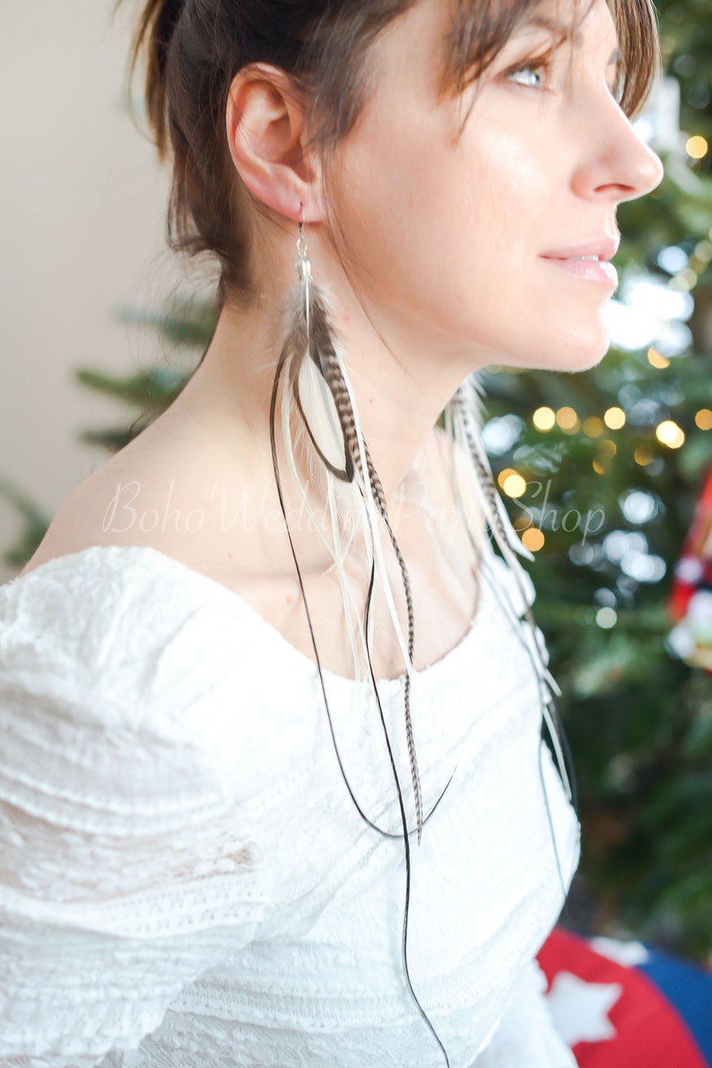 May include: Long feather earrings with a mix of white, brown, and black feathers. The earrings are suspended from silver hooks and dangle gracefully. The model is wearing a white off-the-shoulder top.