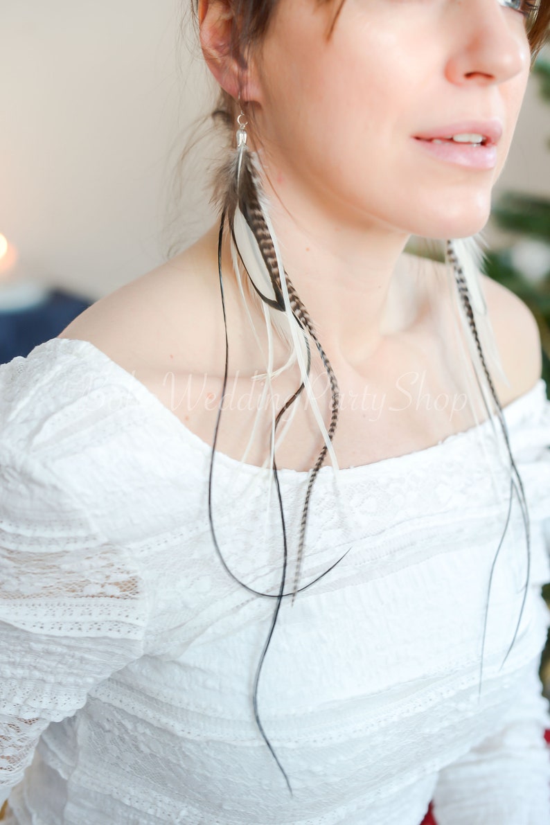May include: Feather earrings with a mix of black, white, and brown feathers. The earrings are long and dangle, with a silver hook. The model is wearing a white, off-the-shoulder lace top. The earrings are a bohemian style.