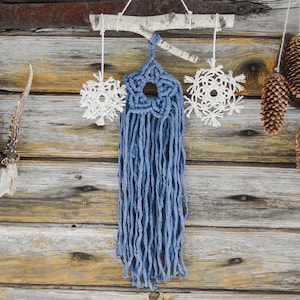 May include: A blue macrame wall hanging with a star design and white crocheted snowflakes on either side. The hanging is displayed on a rustic wooden wall with antlers and pine cones.