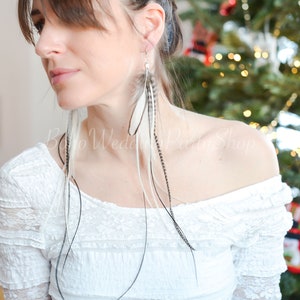 May include: Long feather earrings with white, black, and gray feathers. The earrings are hanging from silver hooks. The model is wearing a white off-the-shoulder top. The background includes a Christmas tree.