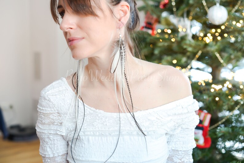 May include: Long feather earrings with white, black, and gray feathers. The earrings are hanging from silver hooks. The model is wearing a white off-the-shoulder top. The background includes a Christmas tree.