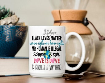 May include: White ceramic coffee mug with a black and blue graphic that reads "I believe... BLACK LIVES MATTER womens rights are human rights NO HUMAN IS ILLEGAL SCIENCE IS REAL LOVE IS LOVE & KINDNESS IS EVERYTHING".