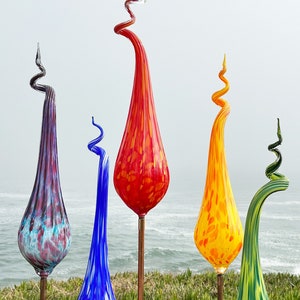 Op de afbeelding: Vijf kleurrijke glazen tuinstokken met een traanvorm en een spiraalvormig ontwerp. De stokken zijn gemaakt van glas en zijn verkrijgbaar in rood, oranje, geel, blauw en groen.