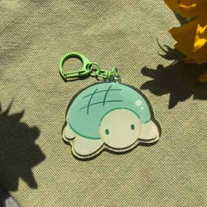 May include: A turtle-shaped keychain with a light green body and a white belly. The turtle has a green grid pattern on its shell and a green clasp. The background is a textured, olive-green fabric.