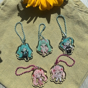 May include: Five acrylic charm keychains featuring anime-style characters with colorful hair and outfits. The keychains have a gold border and are attached to a beaded chain. A yellow sunflower is in the background.
