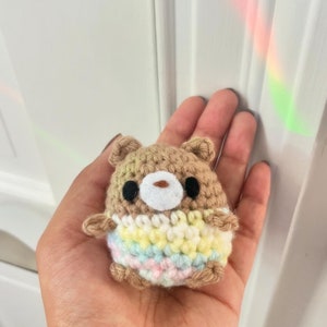 May include: A small, handmade crochet bear in shades of brown, beige, and pastel rainbow colors. The bear has black eyes and a white muzzle. The bear is approximately 2 inches tall.