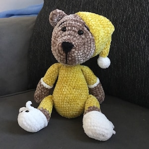 Puede incluir: Un oso de peluche hecho a mano con un pelele y gorro de dormir amarillos. El oso tiene pelaje marrón en la cabeza, los brazos y las piernas, con detalles blancos en los pies y las mangas. El oso está sentado sobre una superficie gris.