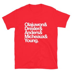 May include: Red short-sleeved t-shirt with white text that reads "Olajuwon & Drexler & Anders & Micheaux & Young."