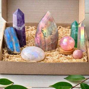 Puede incluir: Una caja de regalo de cartón rellena de virutas de madera contiene una colección de cristales y piedras pulidas. El conjunto incluye una placa de labradorita azul, una punta de amatista morada, una gran piedra iridiscente en forma de llama, una piedra lunar lisa, una esfera rosa, una punta verde, una punta alta iridiscente y una pequeña esfera rosa.