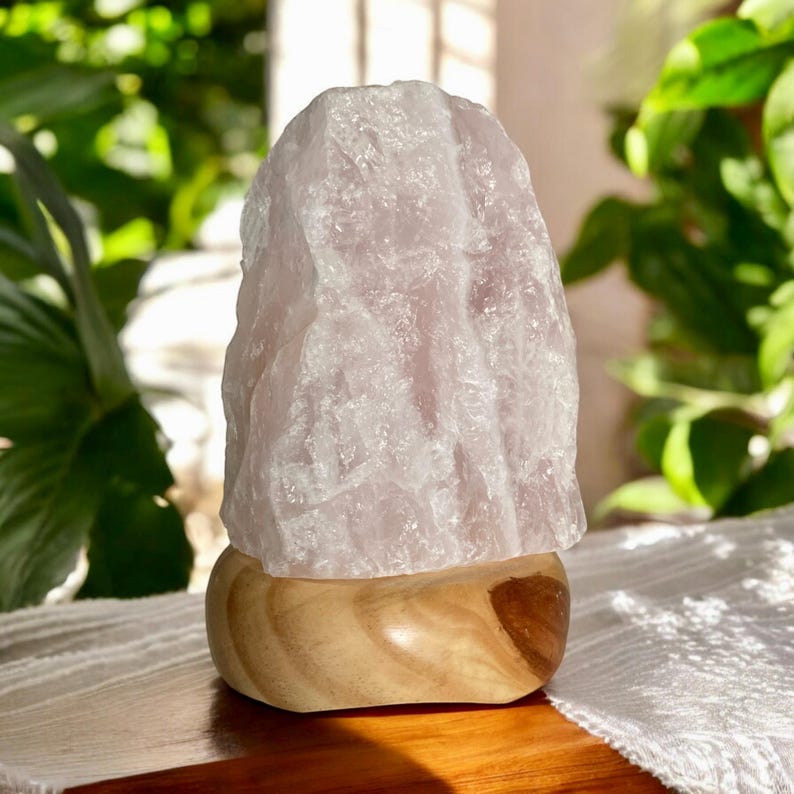 May include: A large, roughly shaped rose quartz crystal lamp sits on a smooth, light-coloured wooden base. The lamp emits a soft, pink glow, and is surrounded by green foliage. The lamp is a decorative item.