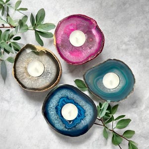 May include: Four circular agate stone candle holders, each featuring a central white tea light candle. The stones are dyed in vibrant shades of magenta, teal, dark blue, and a natural translucent gray, all with distinct crystalline centers.