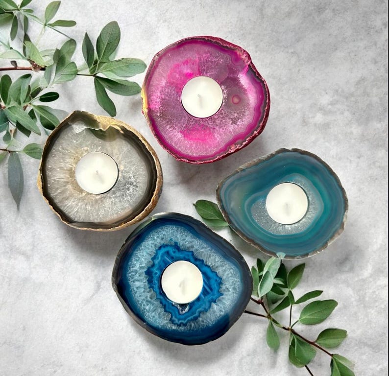 May include: Four circular agate stone candle holders, each featuring a central white tea light candle. The stones are dyed in vibrant shades of magenta, teal, dark blue, and a natural translucent gray, all with distinct crystalline centers.