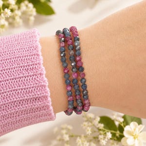 May include: A triple-strand bracelet composed of small, faceted, round beads in alternating shades of deep blue and vibrant magenta. The beads are strung together in a repeating pattern, creating a textured appearance on the wrist.