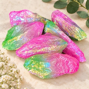 May include: A collection of raw, irregularly shaped crystals featuring a vibrant iridescent coating. The stones display a gradient of bright pink and lime green hues with a metallic, rainbow-like sheen across their textured, jagged surfaces.