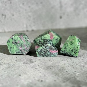 May include: Four faceted, raw ruby in zoisite stones. The stones are a mix of green, black, and pink. The stones are arranged on a gray surface. The stones have a rough, natural texture.