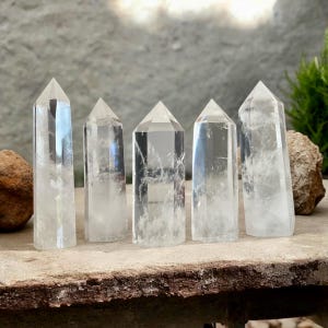 Puede incluir: Cinco puntas de cristal de cuarzo claro, también conocidas como torres de cuarzo claro, están dispuestas en una fila sobre una superficie de piedra rugosa de color marrón. Los cristales son todos de tamaño y forma aproximadamente iguales, y tienen un acabado natural sin pulir.